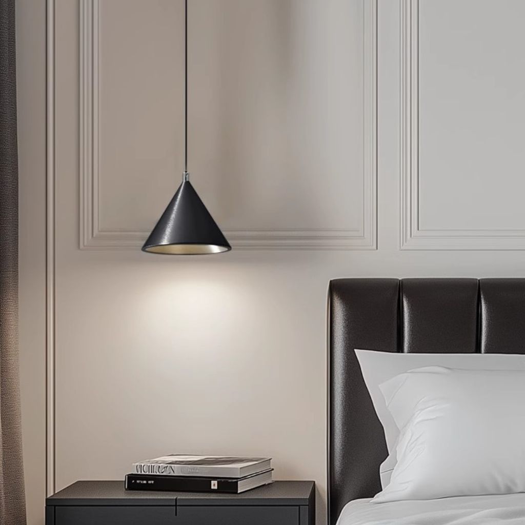 A modern black cone-shaped pendant light hanging above a bedside table in a bedroom setting, casting a warm glow. The scene includes a dark leather headboard, white pillows, and books on the table.