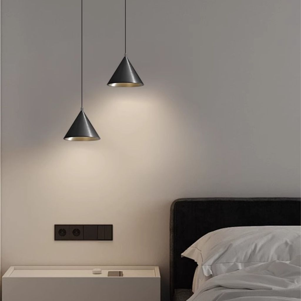 Two modern cone-shaped pendant lights in a dark gray finish with a brushed gold interior, hanging in a minimalist bedroom setting above a bed with white bedding.
