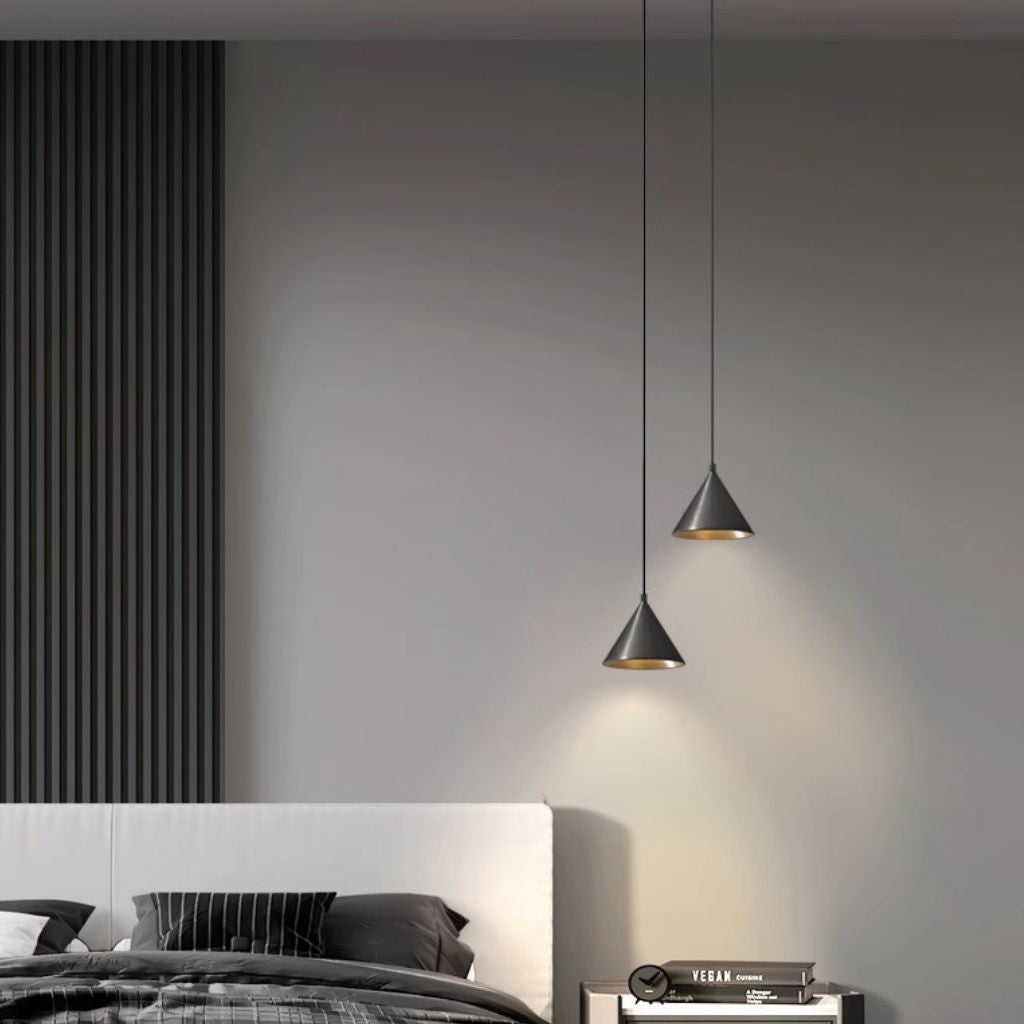 Two modern black cone pendant lights with gold interior linings hanging in a minimalist bedroom setting with a gray wall, white headboard, and dark bedding.