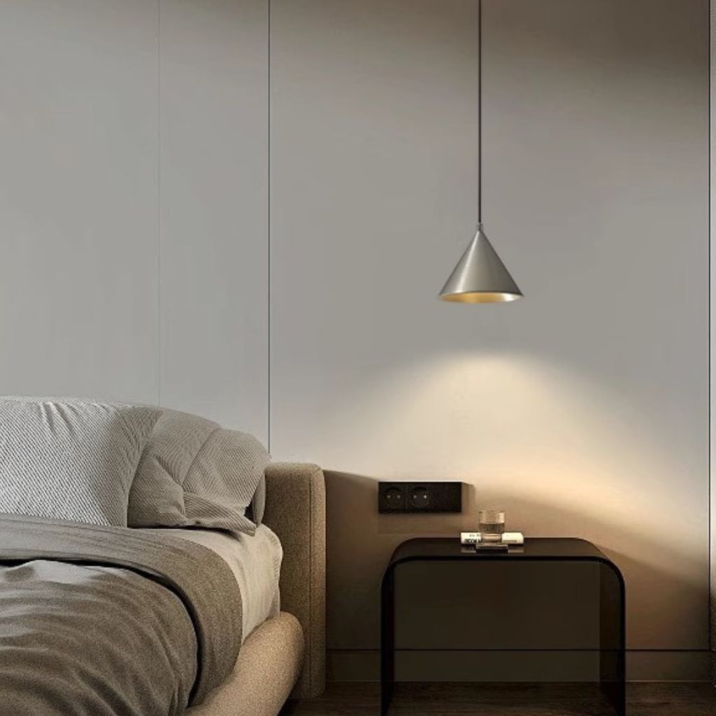 A modern bedroom scene featuring a sleek, cone-shaped pendant light with a brushed silver metal finish and a warm gold interior, hanging above a minimalist black nightstand.