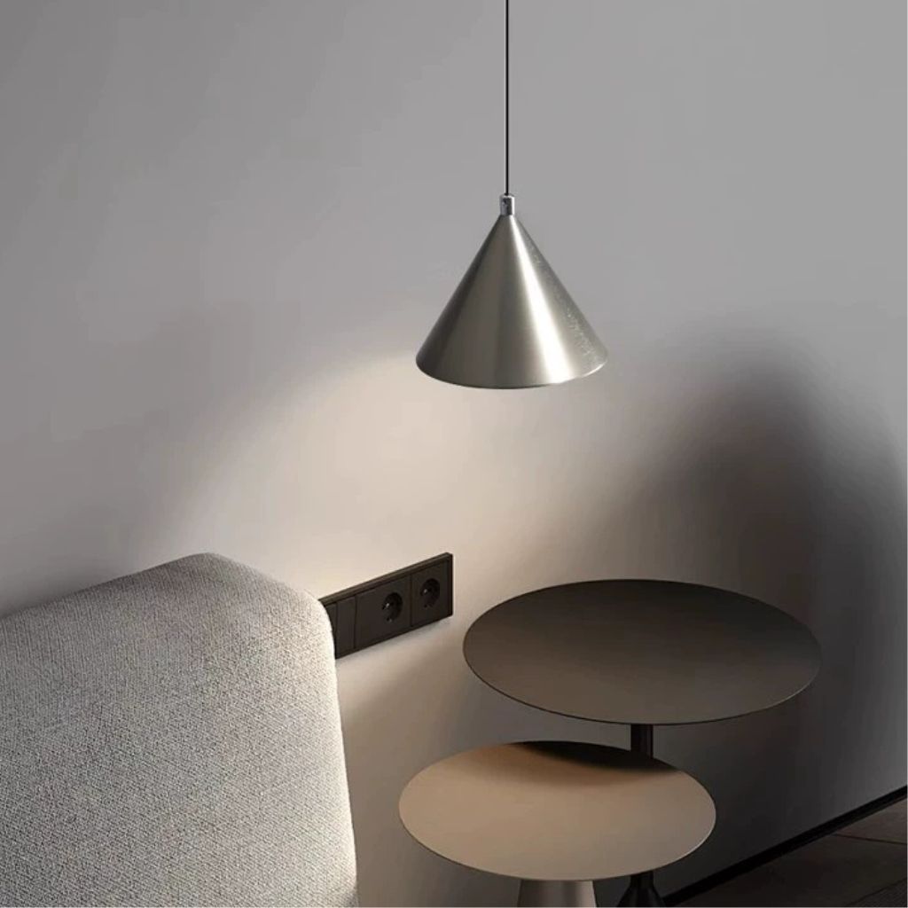 A modern cone-shaped pendant light in brushed silver metal hanging in a minimalist room corner next to a gray upholstered chair and two round nesting side tables.